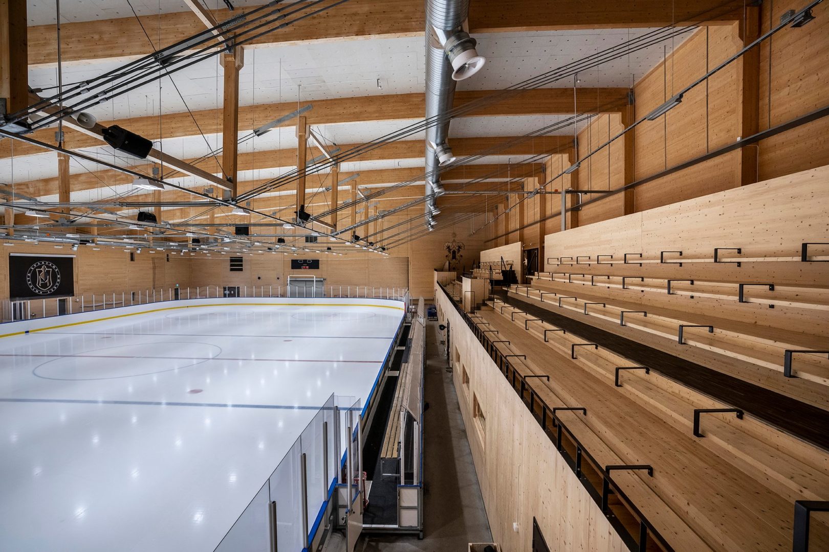 Upplyst inomhus-ishall med tillhörande läktare bestående av sittrader i trä, med svarta armstöd med jämna mellanrum. 