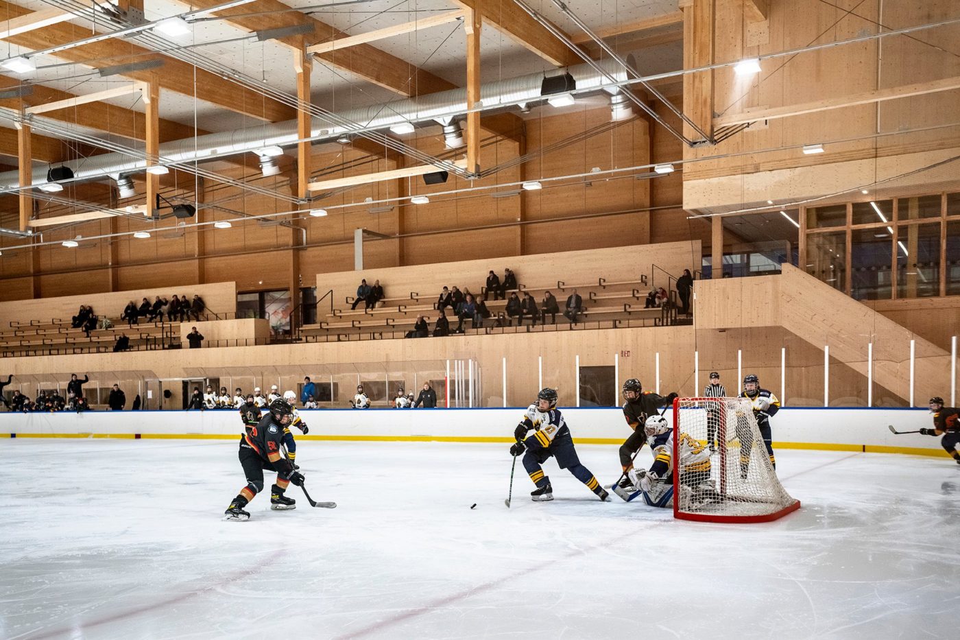 5 stycken hockeyspelare och en målvakt. 3 utespelare framför målburen på väg mot en puck och två utespelare snett bakom målet. Folk syns på håll, sittandes på läktare. 